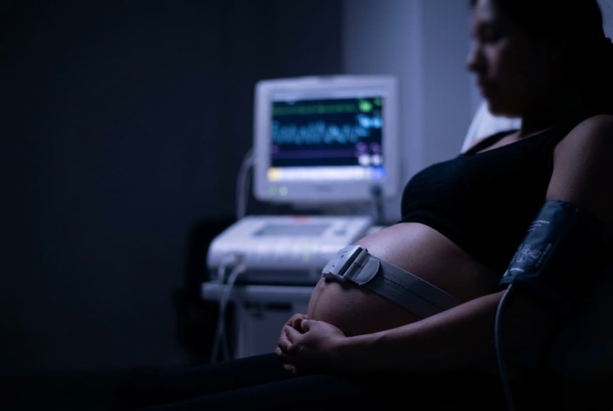 Abdomen de una mujer embarazada con cinturón de monitoreo fetal y brazalete de presión arterial, con el trazo de cardiotocografía en el monitor