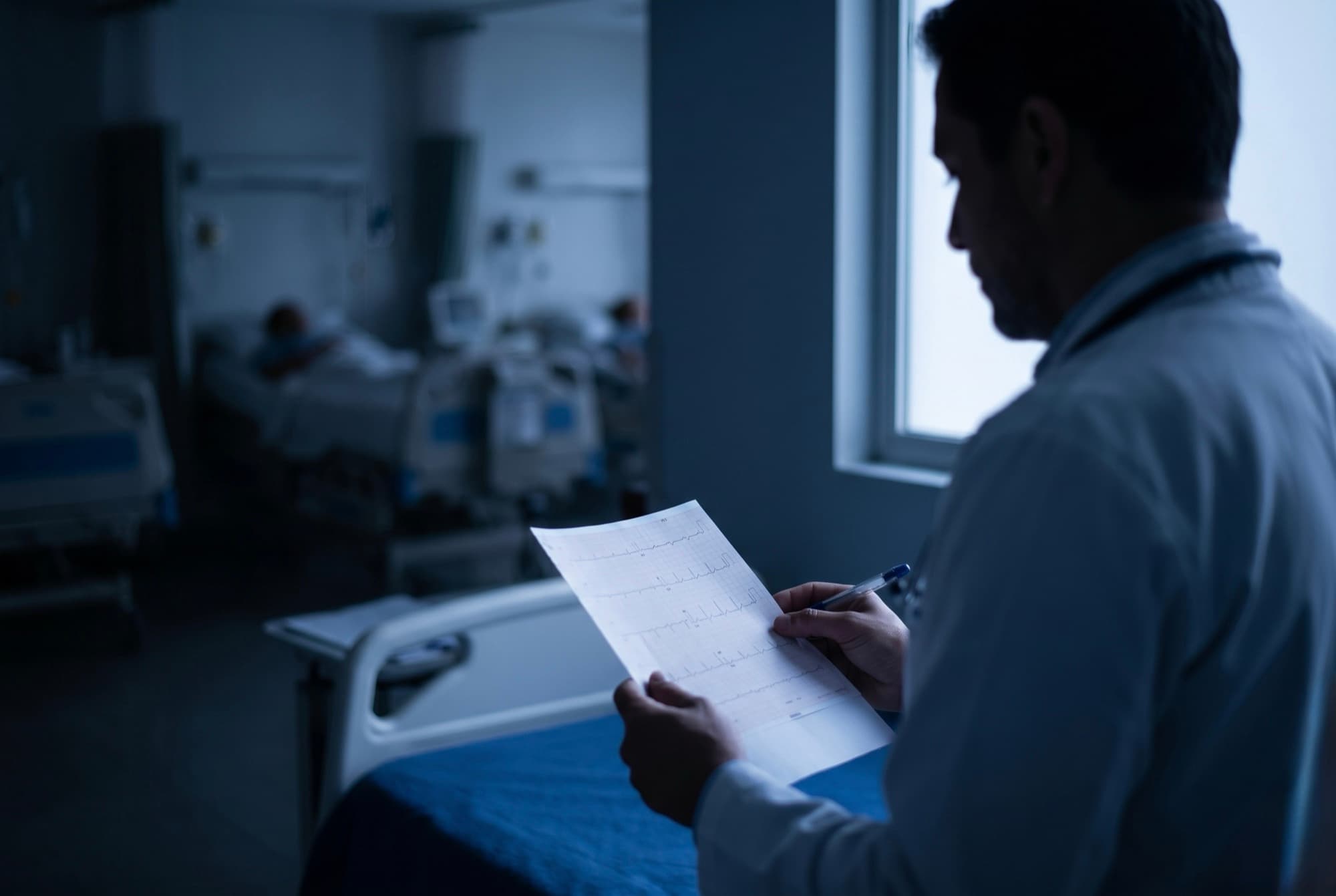 Internista de bata blanca leyendo un electrocardiograma frente a una sala de hospitalizacion