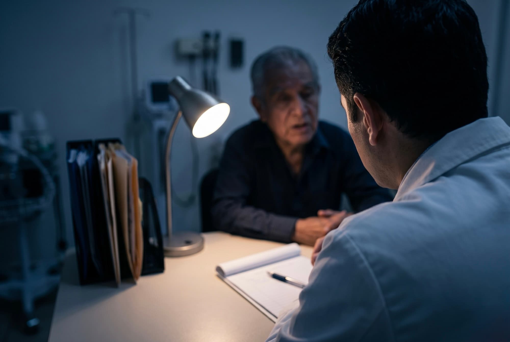 Médico escuchando con atención a un paciente mayor al otro lado del escritorio, con una libreta de notas y sin pantalla de por medio