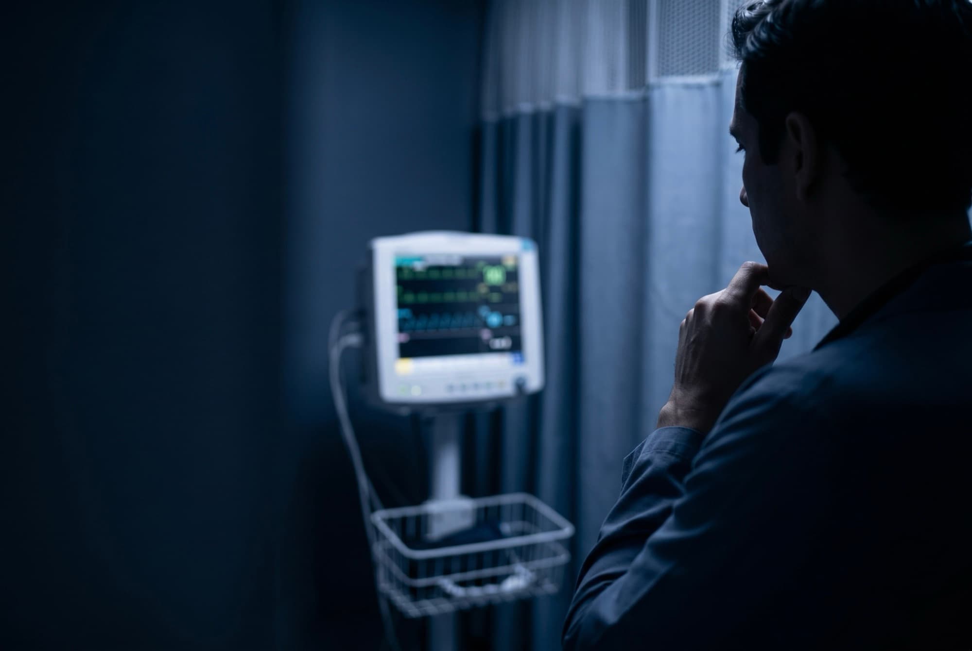 Médico reflexionando frente a un monitor de signos vitales en un box de urgencias