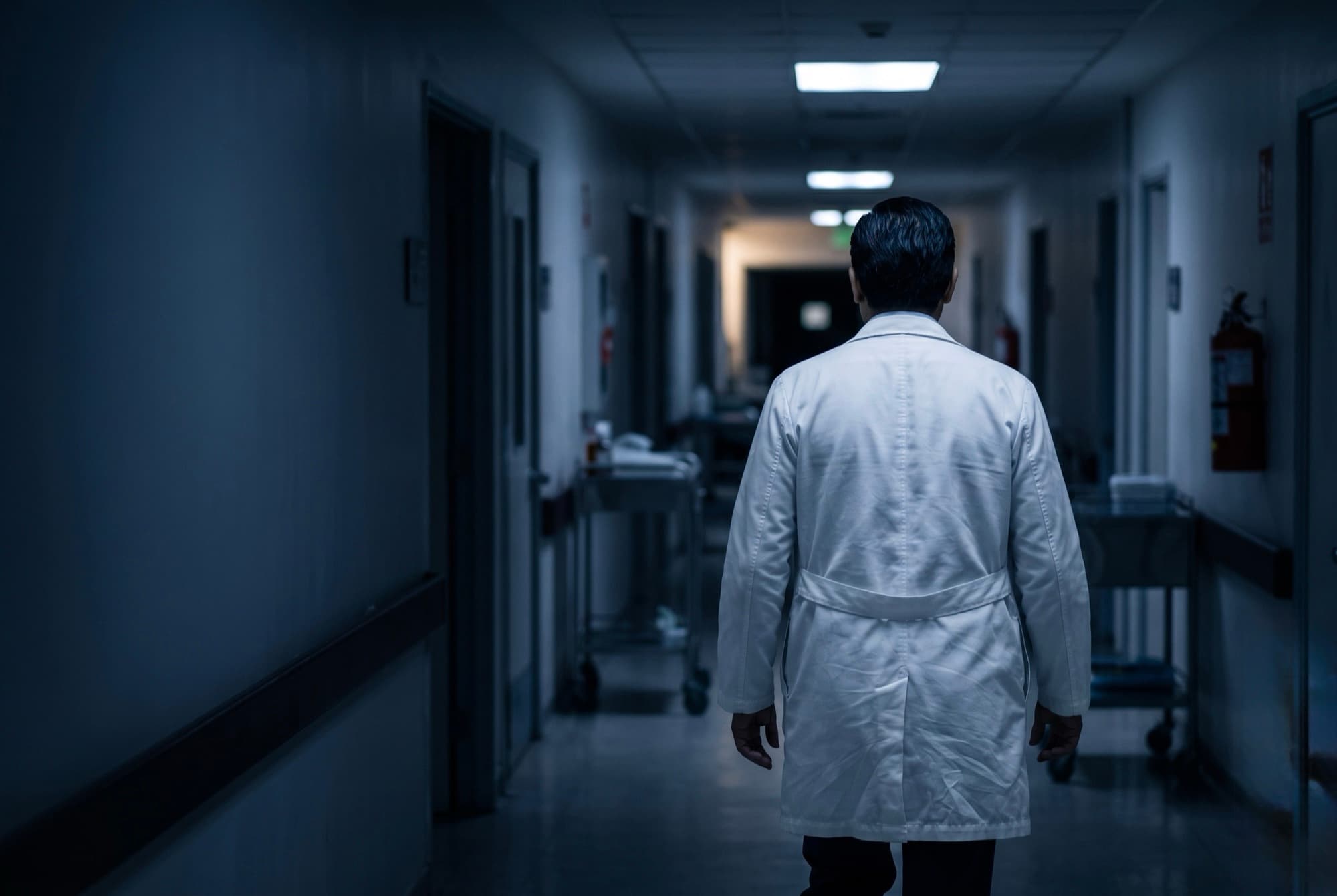 Internista de bata blanca, de espaldas, caminando por un pasillo de hospital en penumbra